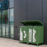 Trois poubelles extérieures vertes étiquetées pour les bouteilles/canettes, les déchets et les produits organiques se trouvent à côté d'un bâtiment moderne aux murs de verre.