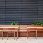 Two brown metal and wood 1050 Series tables with FORO Series chairs are placed in front of a long wooden bench with planters, set against a wall with horizontal gray slats.