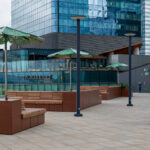 Terrasse extérieure moderne avec des bancs en bois Maglin, des tables et des chaises en métal, des parasols verts et de grands bâtiments en verre comme la tour Stantec en arrière-plan. L'enseigne 