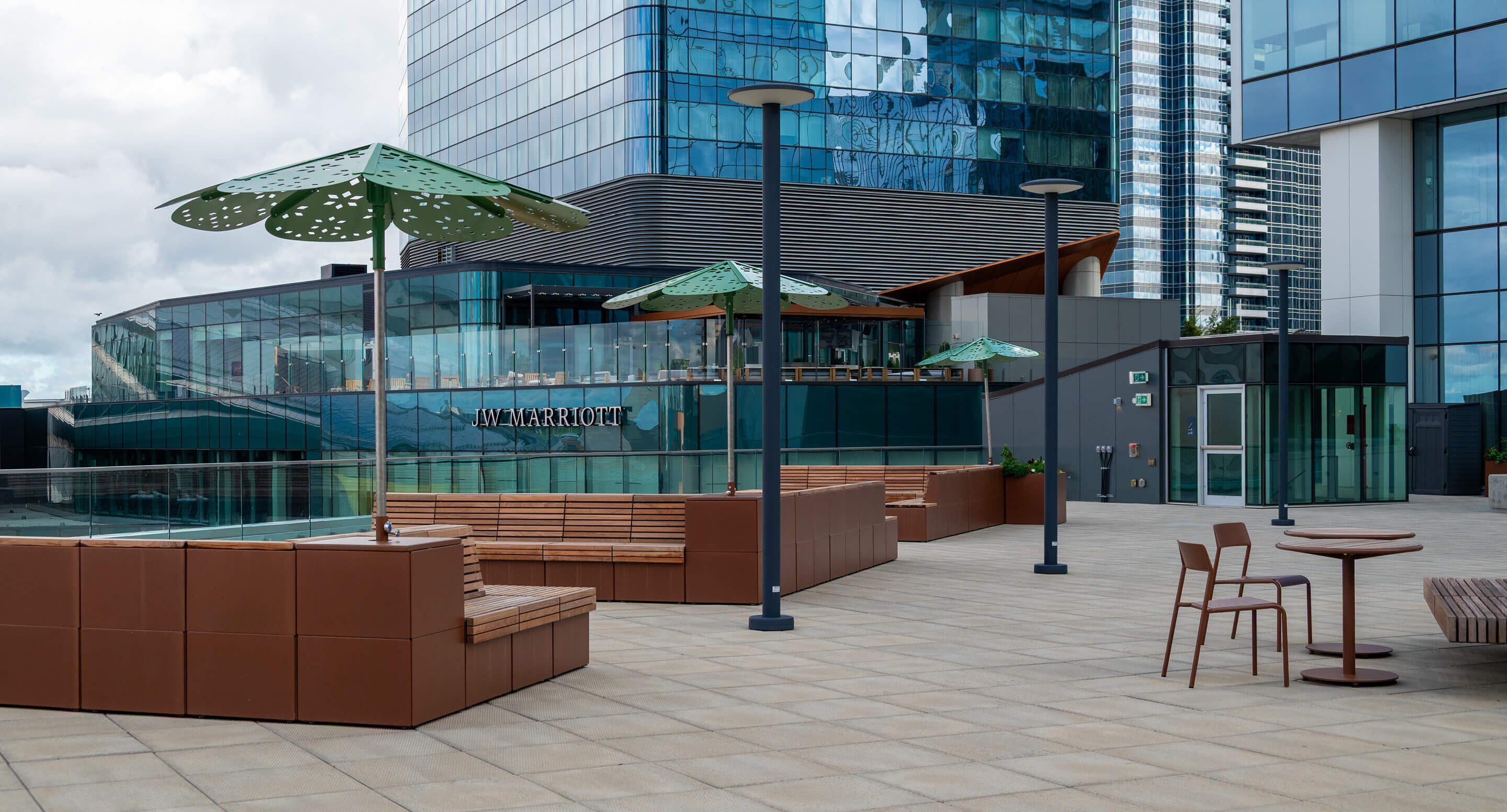 Outdoor terrace with Maglin wooden benches, tables, chairs, and green umbrella shades, situated next to the modern glass Stantec Tower and Marriott signage—an inviting Rooftop Retreat in the city.