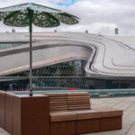 Modern outdoor seating area with wooden Pixel Collection benches and a decorative ROMA Sun Shade umbrella on a terrace, with a curved, futuristic building - Rogers Place Arena - in the background under a cloudy sky.