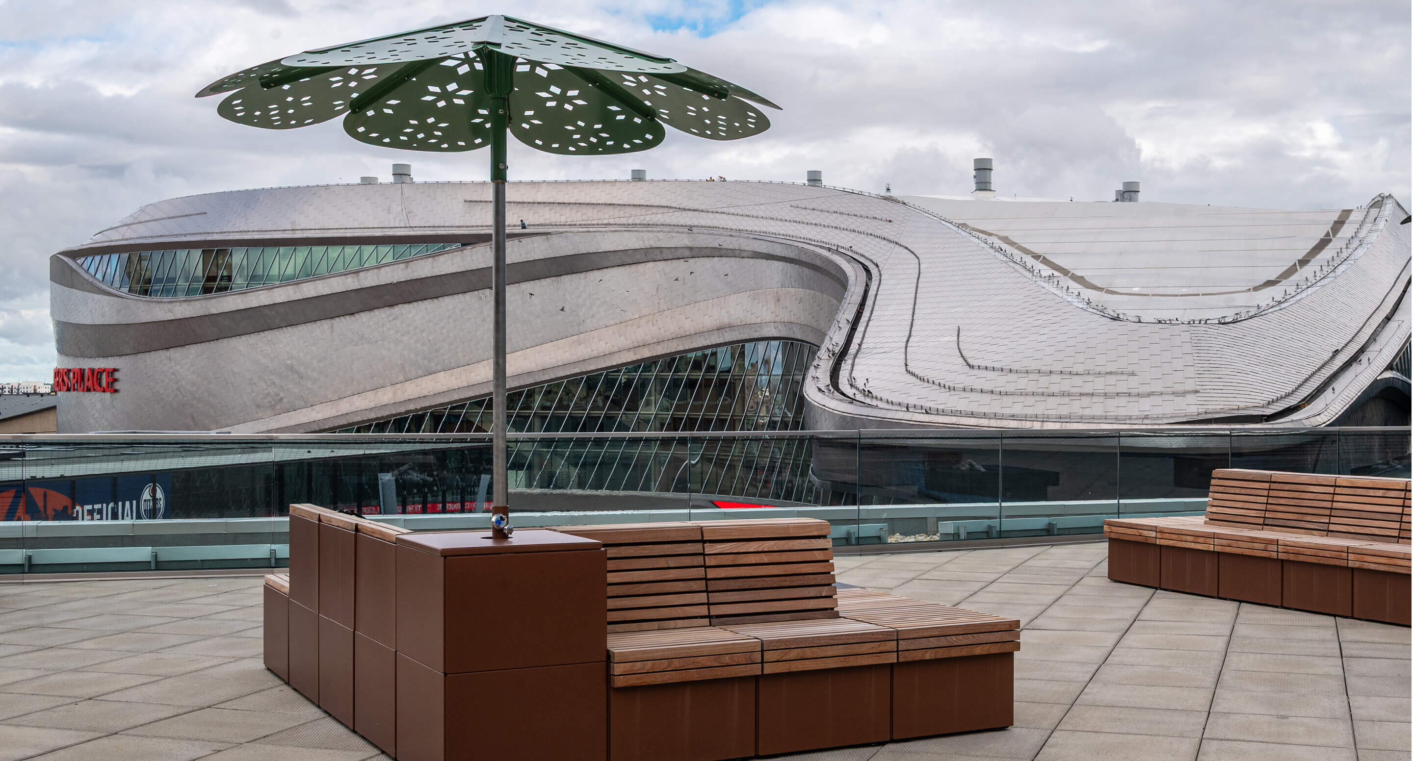 Modern outdoor seating area with wooden Pixel Collection benches and a decorative ROMA Sun Shade umbrella on a terrace, with a curved, futuristic building - Rogers Place Arena - in the background under a cloudy sky.