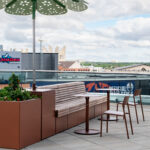 Banquettes et chaises brunes, jardinière avec de la verdure et un parasol décoratif sur un patio sur le toit avec vue sur les bâtiments de la ville.