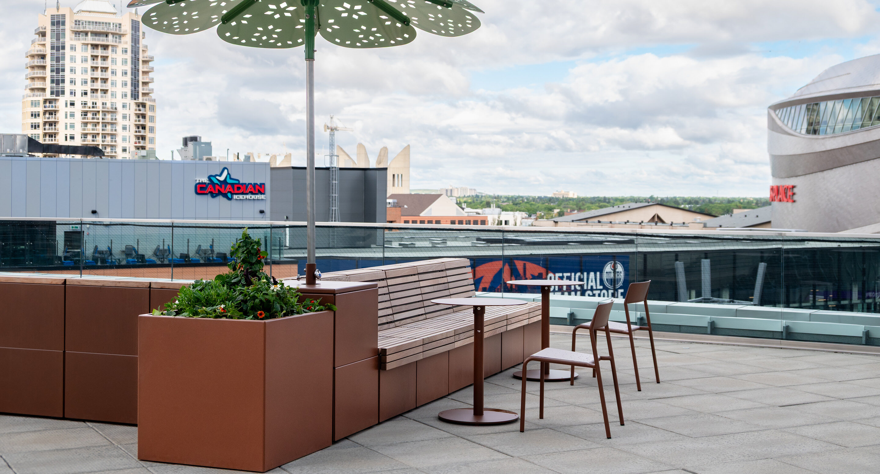 Banquettes et chaises brunes, jardinière avec de la verdure et un parasol décoratif sur un patio sur le toit avec vue sur les bâtiments de la ville.