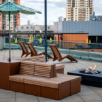 Terrasse sur le toit avec des bancs en bois, des chaises longues, un parasol vert, un foyer moderne et des bâtiments de la ville en arrière-plan.