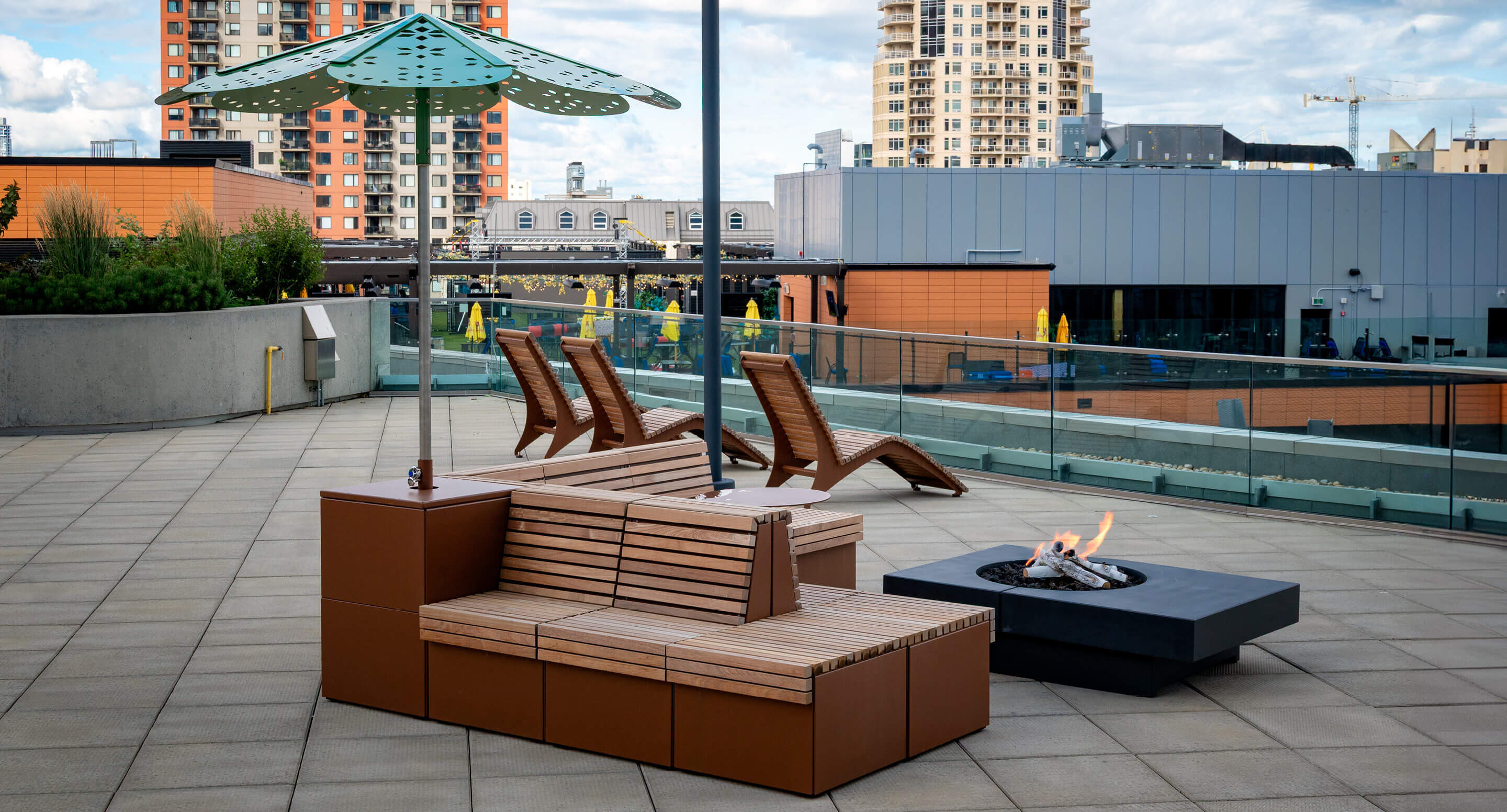 Terrasse sur le toit avec des bancs en bois, des chaises longues, un parasol vert, un foyer moderne et des bâtiments de la ville en arrière-plan.