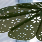Close-up of the ROMA Sun Shade - a green metal structure with petal-shaped panels featuring geometric cutouts, resembling a stylized flower, set against a blurred urban background.