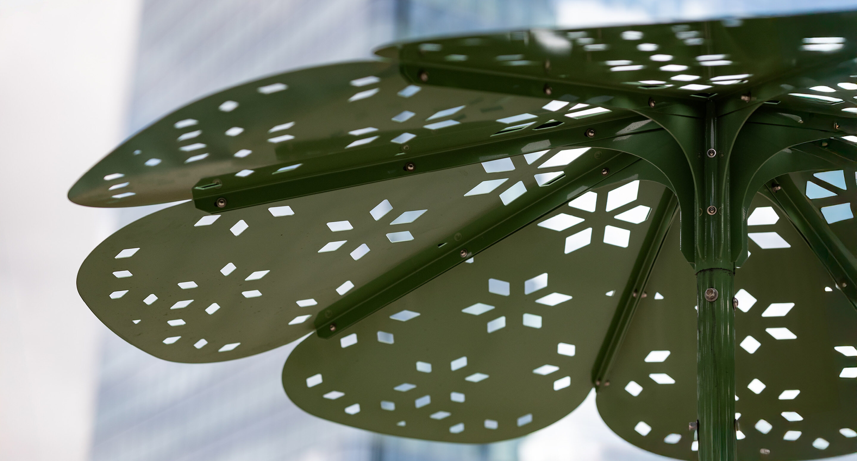 stantec-tower-roma-sun-shade-detail Gros plan d'un auvent vert en métal en forme de fleur avec des découpes en forme de flocons de neige, sur un fond flou de grands immeubles en verre.