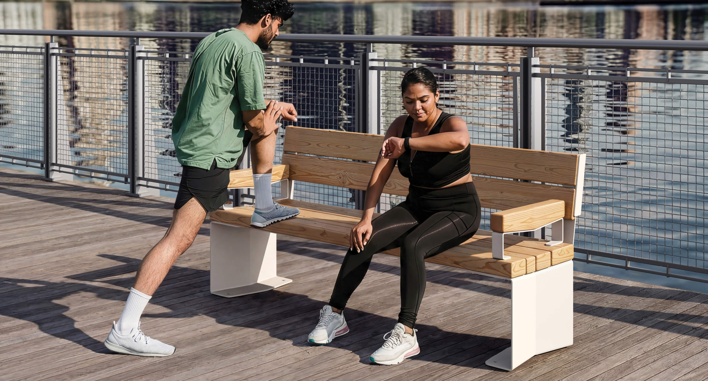 Un homme étire sa jambe sur un banc tandis qu'une femme en tenue de sport, rappelant l'élégance sportive de Corolle, est assise et regarde sa smartwatch. Ils sont en plein air, près d'un plan d'eau, avec une clôture métallique en arrière-plan.