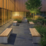 Une cour moderne avec des bancs en bois, un sol en dalles de pierre, des plantes vertes et un arbre - adjacente au bâtiment de Trois avec de grandes baies vitrées et un éclairage intérieur chaleureux ; un espace accueillant reconnu par les prix A+.