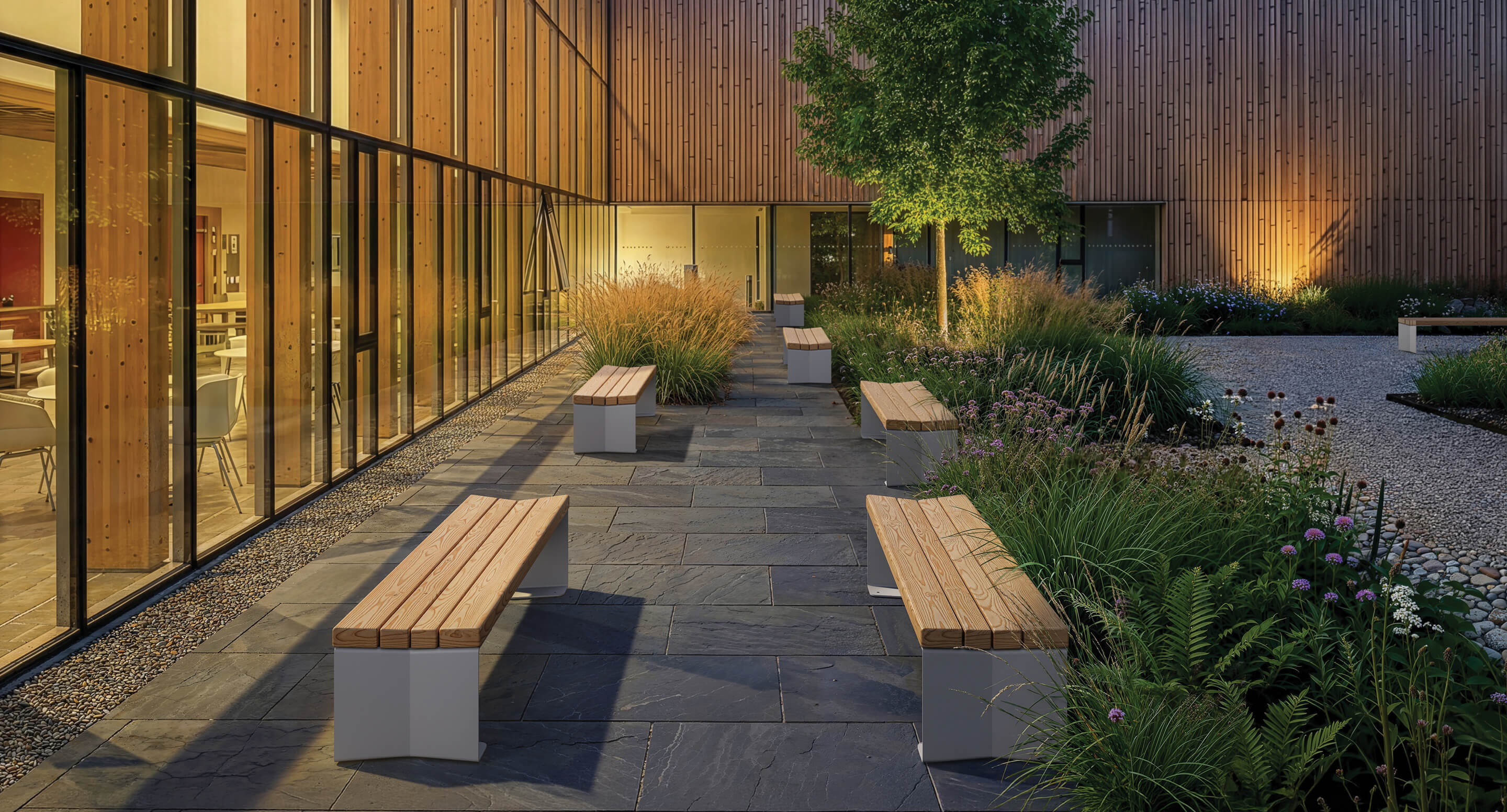 Une cour moderne avec des bancs en bois, un sol en dalles de pierre, des plantes vertes et un arbre - adjacente au bâtiment de Trois avec de grandes baies vitrées et un éclairage intérieur chaleureux ; un espace accueillant reconnu par les prix A+.