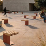 Par une journée ensoleillée, plusieurs personnes se promènent ou s'assoient sur une place extérieure moderne avec des bancs en bois et en métal éparpillés, profitant de la conception primée et reconnue par les prix A+.