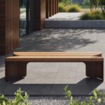 Un banc moderne en bois est posé sur un patio carrelé à l'extérieur d'un bâtiment avec des panneaux en bois, entouré de plantes et de verdure - un endroit accueillant qui rappelle les designs récompensés par les prix A+, parfait pour se détendre ou présenter de charmants jouets Corolle.