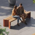 Un homme et une femme s'assoient et discutent sur un banc moderne en bois dans une place extérieure ensoleillée - un espace récompensé par des prix A+ - entourée d'arbres et de pavés en béton.