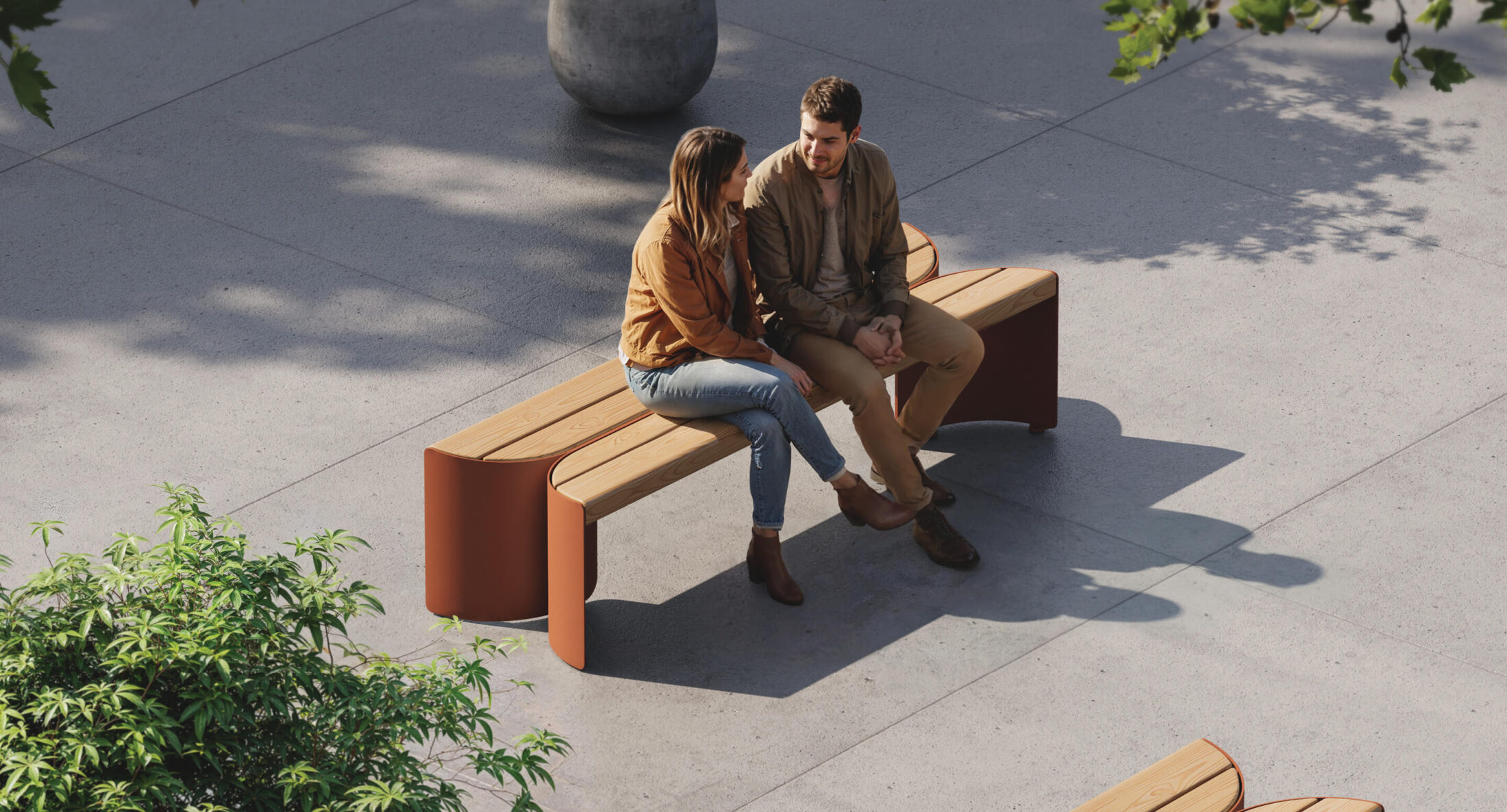 Un homme et une femme s'assoient et discutent sur un banc moderne en bois dans une place extérieure ensoleillée - un espace récompensé par des prix A+ - entourée d'arbres et de pavés en béton.