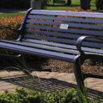Black metal park bench on a paved path, surrounded by green shrubs and red flowers, with a small dedication plaque on the backrest.