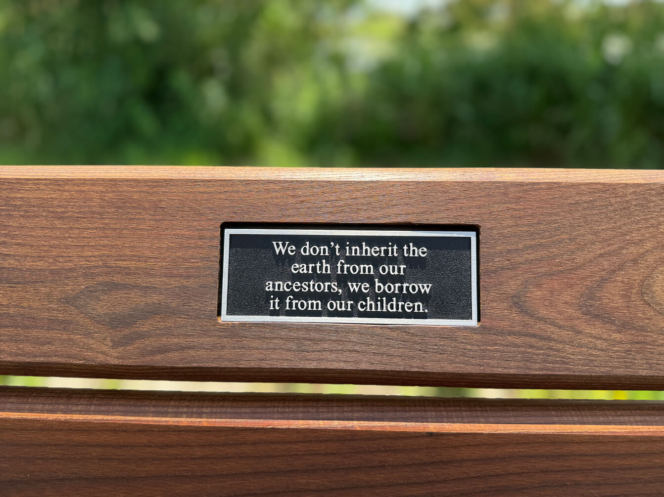 Plaque on a wooden bench reads: "We don’t inherit the earth from our ancestors, we borrow it from our children." Green foliage is blurred in the background.