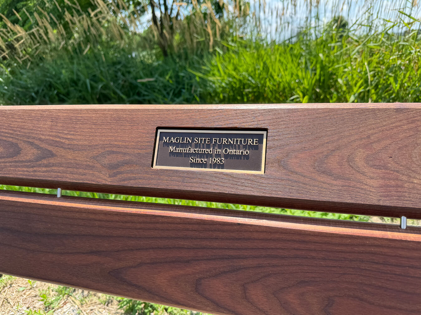 Close-up of a bronze plaque on a wooden bench that reads, "MAGLIN SITE FURNITURE Manufactured in Ontario Since 1983." Grass and trees are visible in the background.