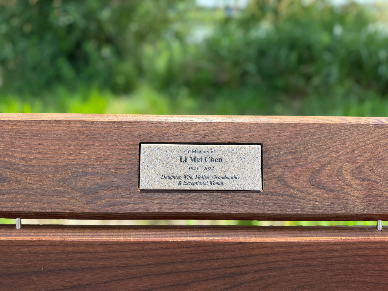 A wooden bench with a plaque that reads: "In Memory of Li Mei Chen 1945-2022 Daughter, Wife, Mother, Grandmother, & Exceptional Woman.