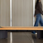 Une personne passe devant un banc moderne en bois avec des pieds noirs devant un mur à lattes verticales.
