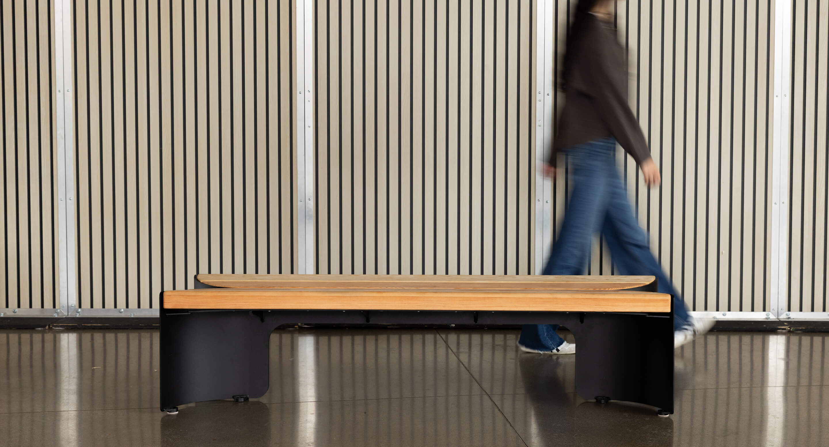 Une personne passe devant un banc moderne en bois avec des pieds noirs devant un mur à lattes verticales.