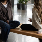 Deux femmes sont assises les jambes croisées sur un banc en bois à l'intérieur, se faisant face et discutant, avec des plantes en pot visibles à l'arrière-plan.