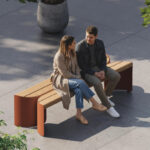 Two people sit on a modern wooden and metal bench outdoors, facing each other and talking, with plants and shadows around them on a paved surface.