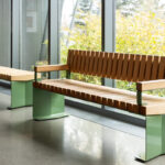 Deux bancs modernes en bois avec des pieds en métal vert sont placés le long d'une grande fenêtre, avec vue sur la verdure à l'extérieur.