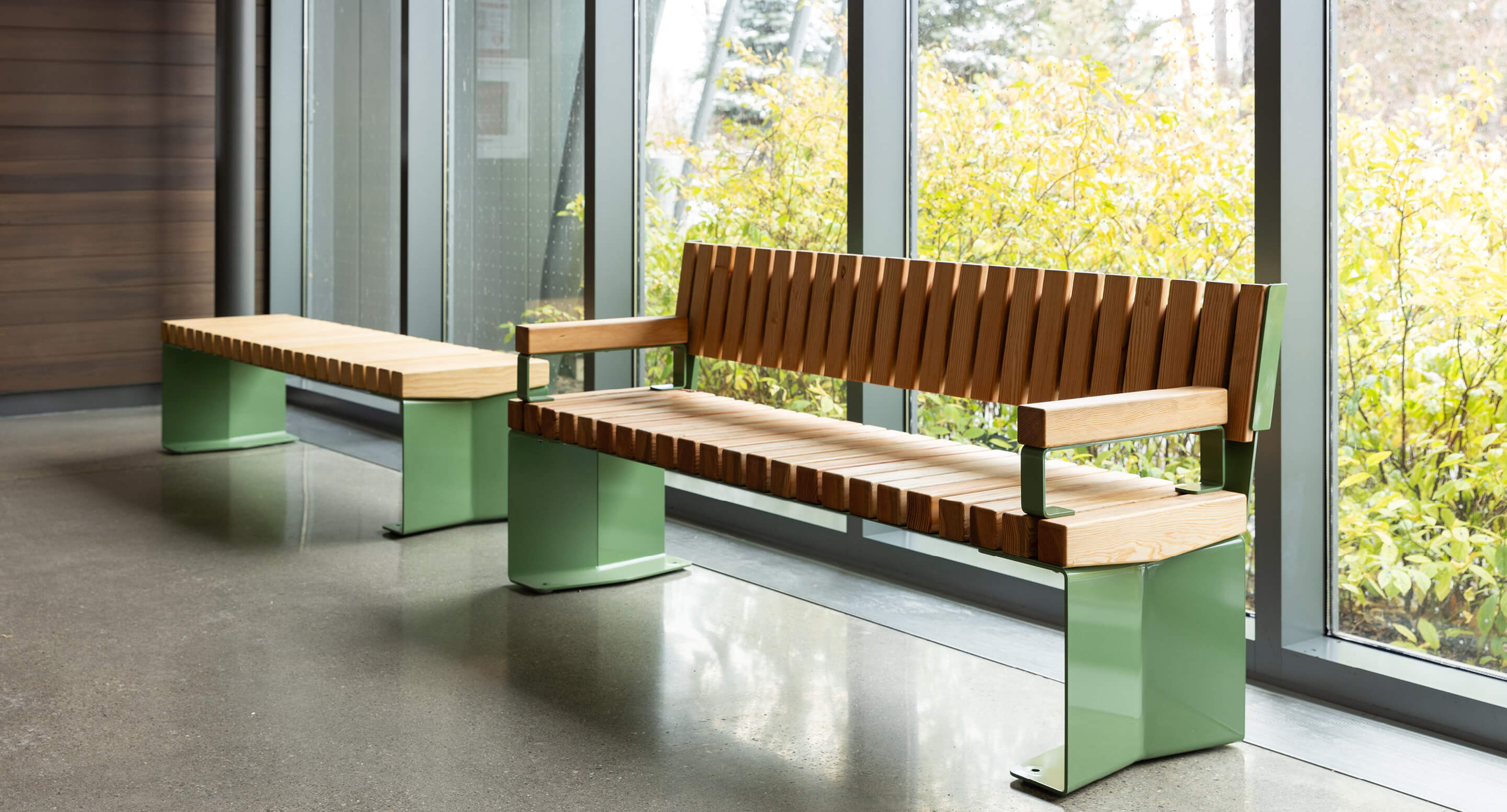 Deux bancs modernes en bois avec des pieds en métal vert sont placés le long d'une grande fenêtre, avec vue sur la verdure à l'extérieur.