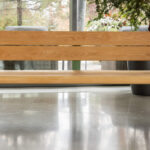 Un banc en bois avec des pieds en métal vert est placé à l'intérieur sur un sol en béton poli, près de grandes fenêtres et de plantes en pot.