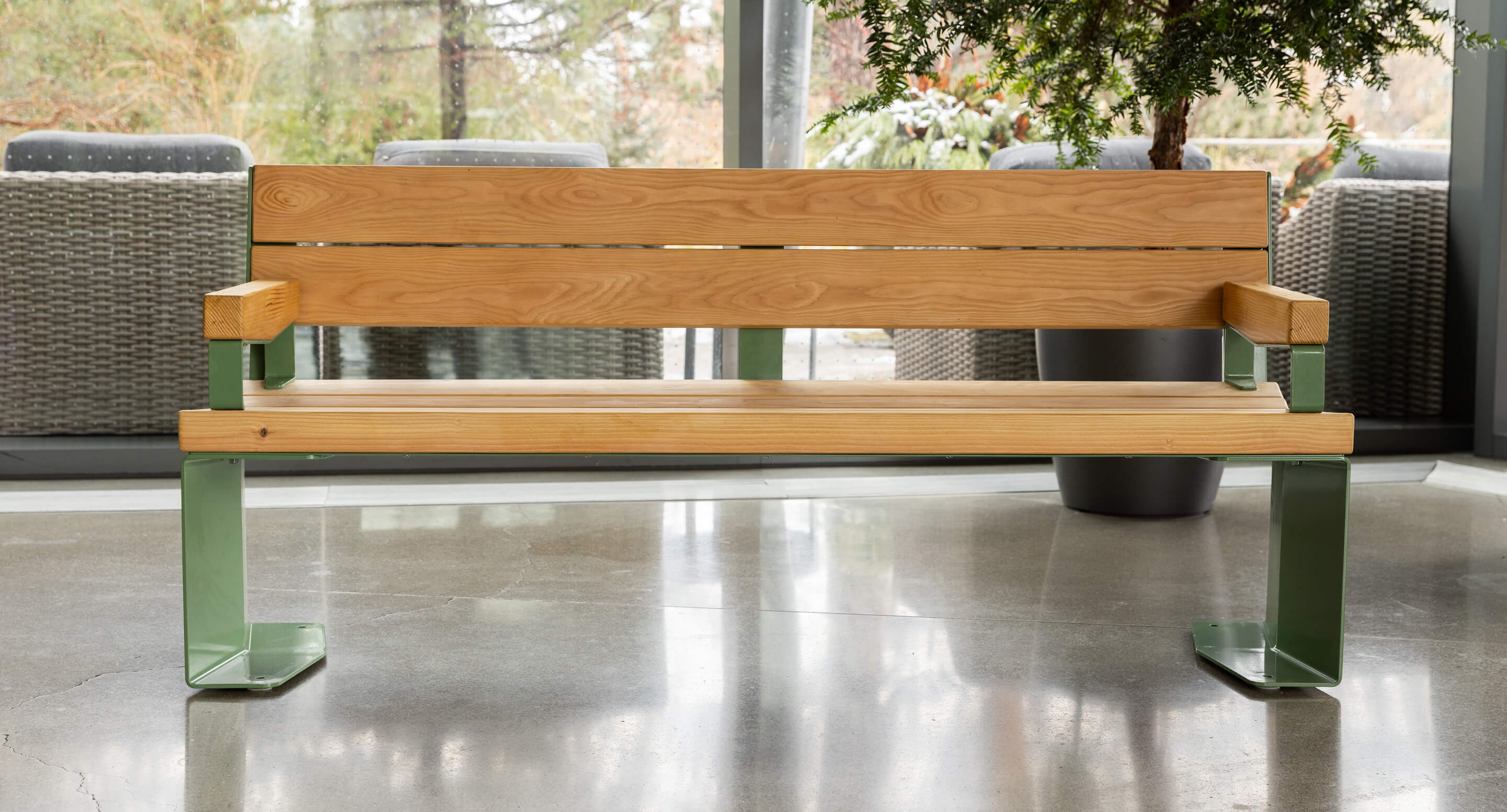 Un banc en bois avec des pieds en métal vert est placé à l'intérieur sur un sol en béton poli, près de grandes fenêtres et de plantes en pot.