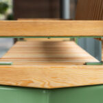 Vue rapprochée d'un banc en bois avec des accoudoirs et des pieds en métal vert, placé à l'intérieur près de grandes fenêtres.