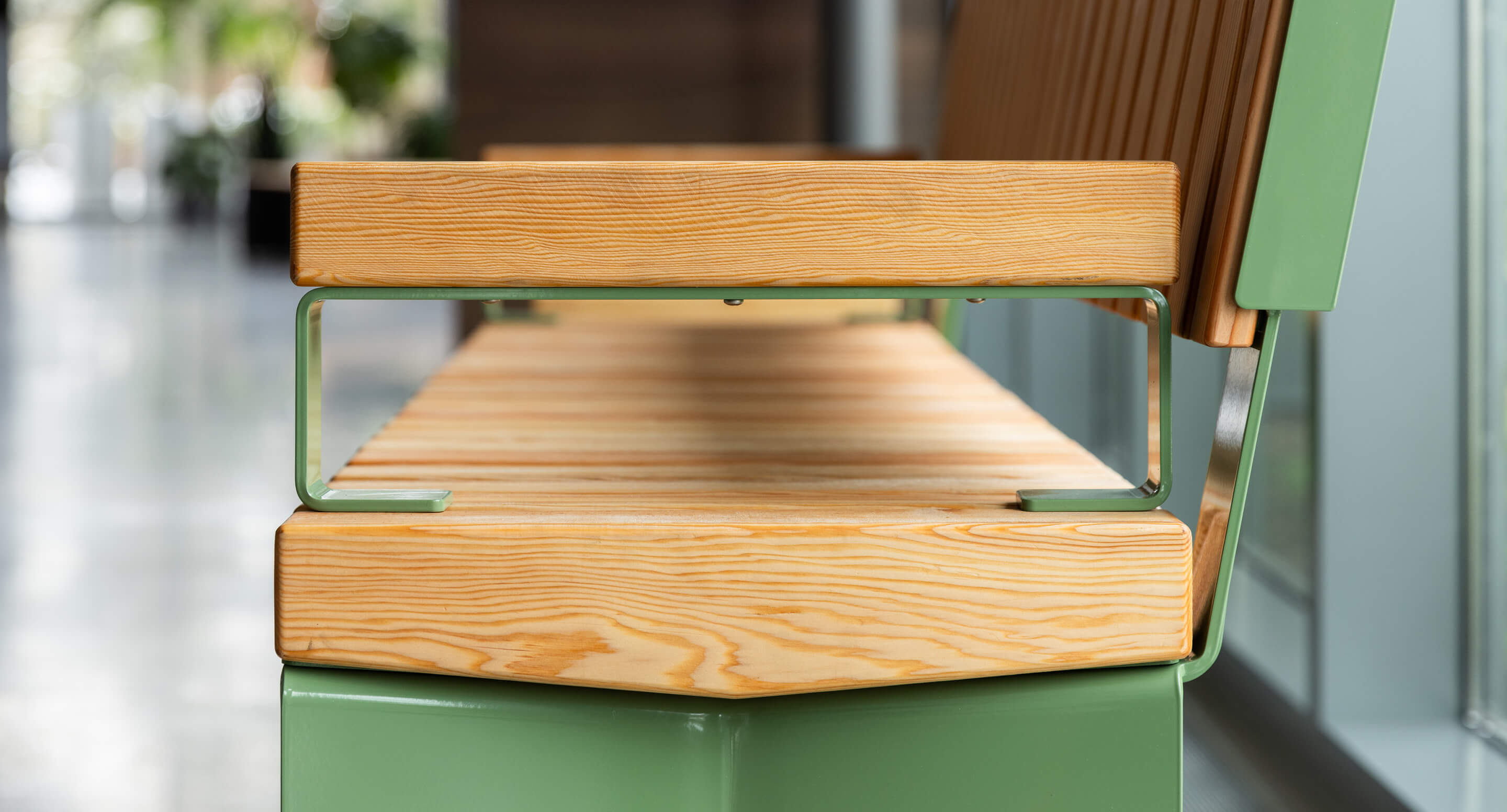 A close-up view of a modern bench with light wood slats and green metal armrests, situated indoors by large windows.