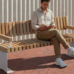 A man sits alone on a wooden bench outdoors, reading a book, with folded legs, on a red brick surface next to a corrugated white wall.