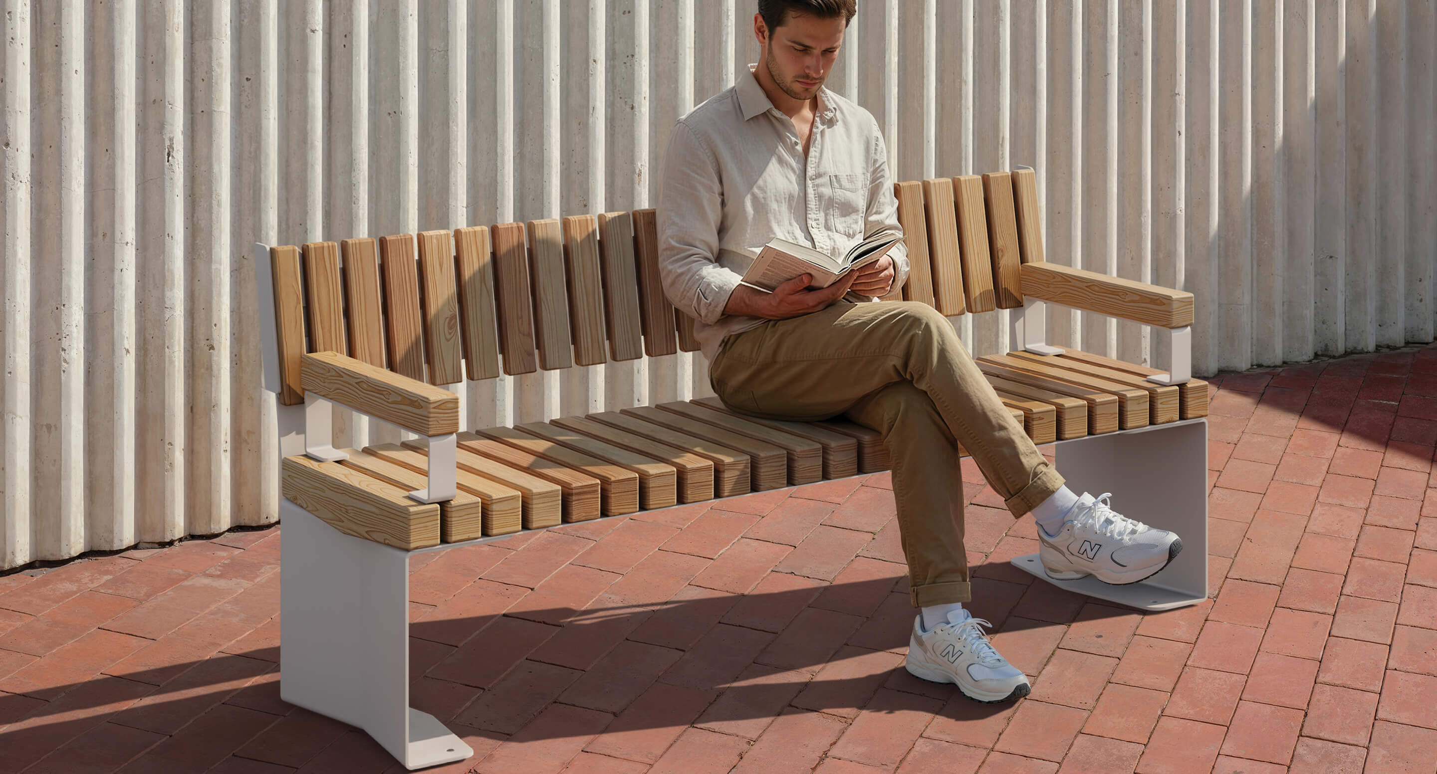 maglin_blox-backed-benches_04 A man sits alone on a wooden bench outdoors, reading a book, with folded legs, on a red brick surface next to a corrugated white wall.
