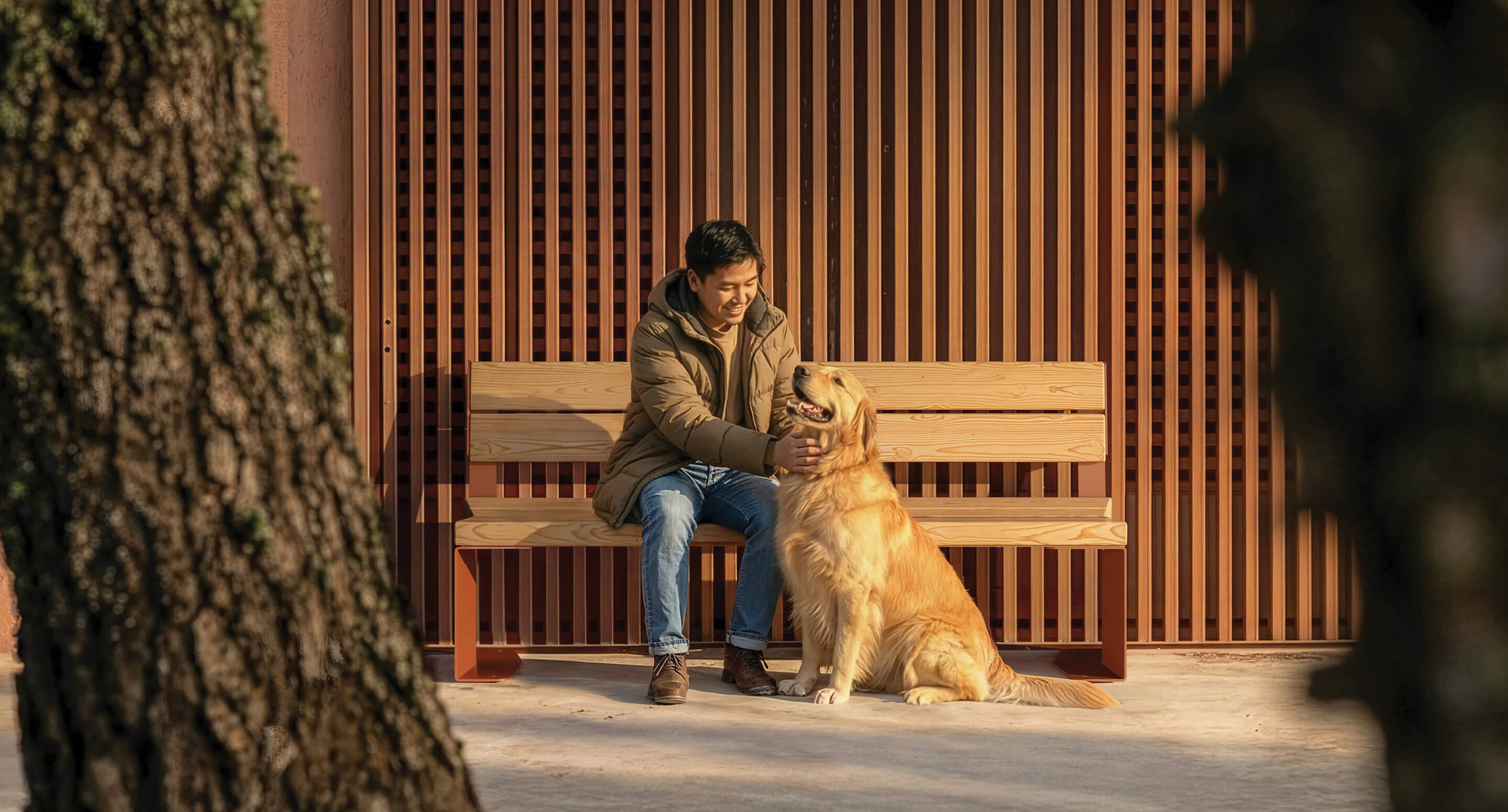 maglin_blox-backed-benches_05 A man in a winter coat sits on a wooden bench outdoors, petting a golden retriever dog, with sunlight casting shadows through a slatted wooden wall behind them.
