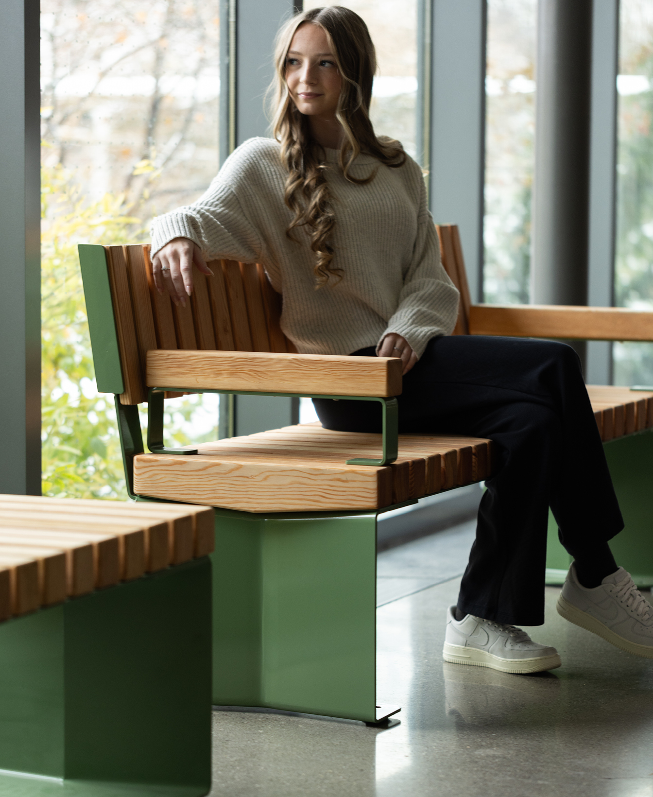 Une femme aux cheveux longs et ondulés est assise sur un banc en bois à l'intérieur, vêtue d'un pull blanc, d'un pantalon sombre et de baskets blanches. De grandes fenêtres laissent entrer la lumière naturelle.