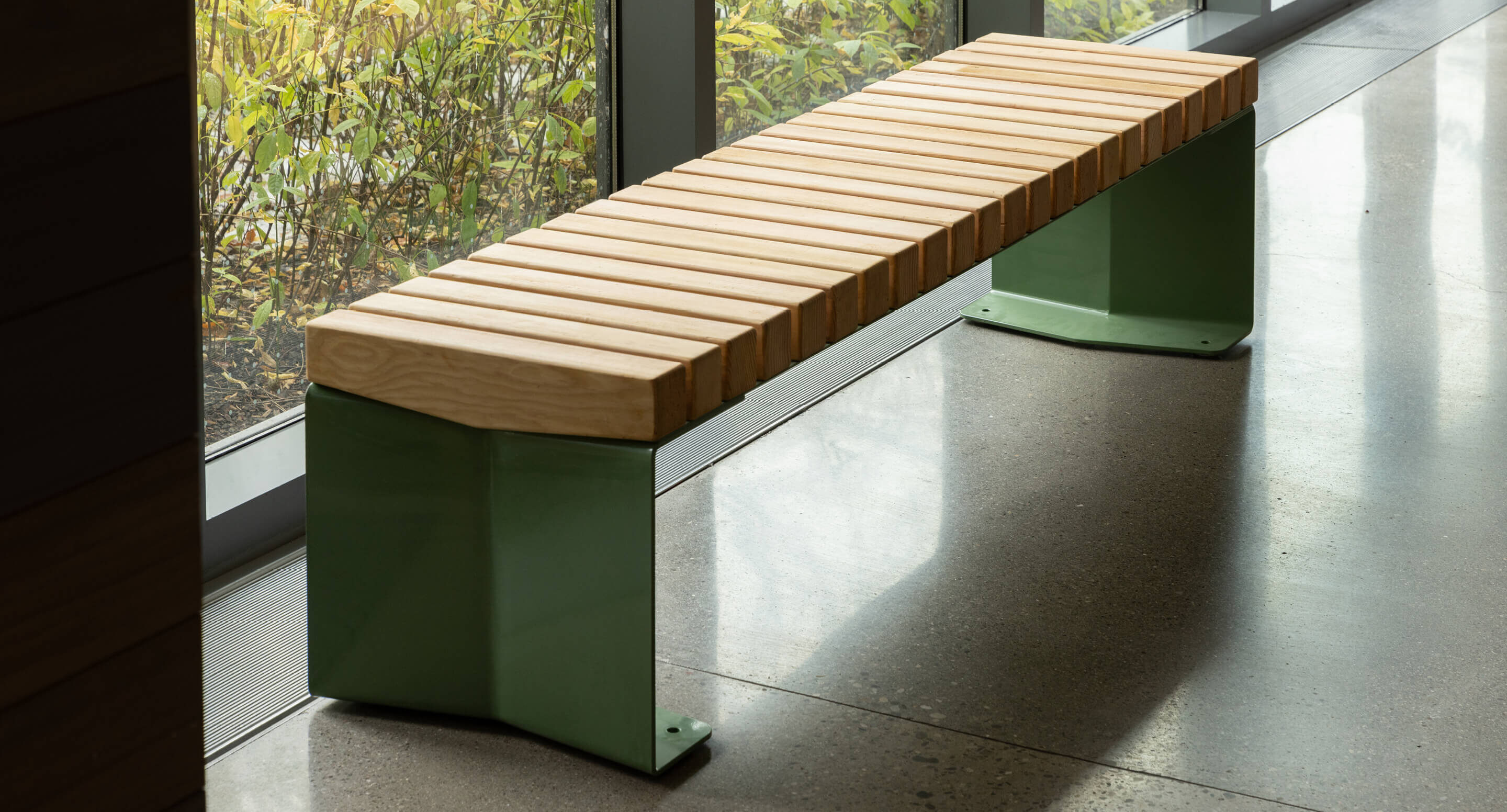 Un banc d'intérieur moderne avec des lattes en bois clair et des pieds en métal vert, placé à côté de grandes fenêtres sur un sol en béton poli.
