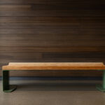 Un banc en bois avec des pieds en métal vert est posé sur un sol poli entre de grandes fenêtres et un mur lambrissé de bois sombre.