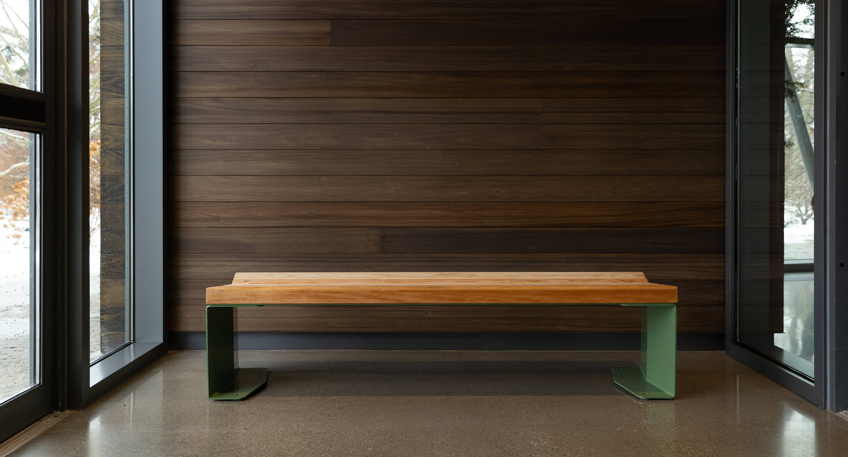 Un banc en bois avec des pieds en métal vert est posé sur un sol poli entre de grandes fenêtres et un mur lambrissé de bois sombre.