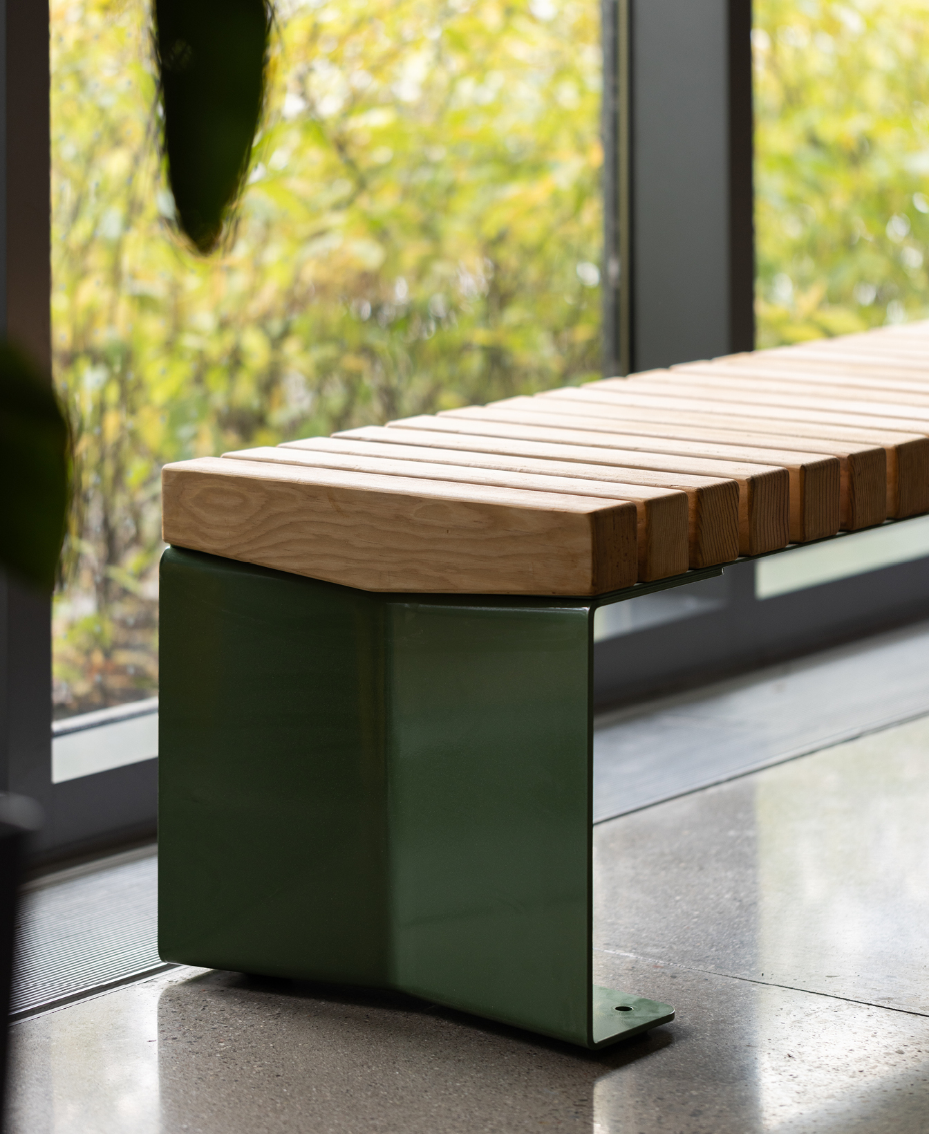 Un banc moderne avec une assise en lattes de bois et une base en métal vert est placé à l'intérieur près de grandes fenêtres avec de la verdure visible à l'extérieur.