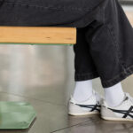 Personne assise sur un banc vert en métal et en bois, vêtue d'un pantalon noir, de chaussettes blanches et de baskets blanches à rayures noires.