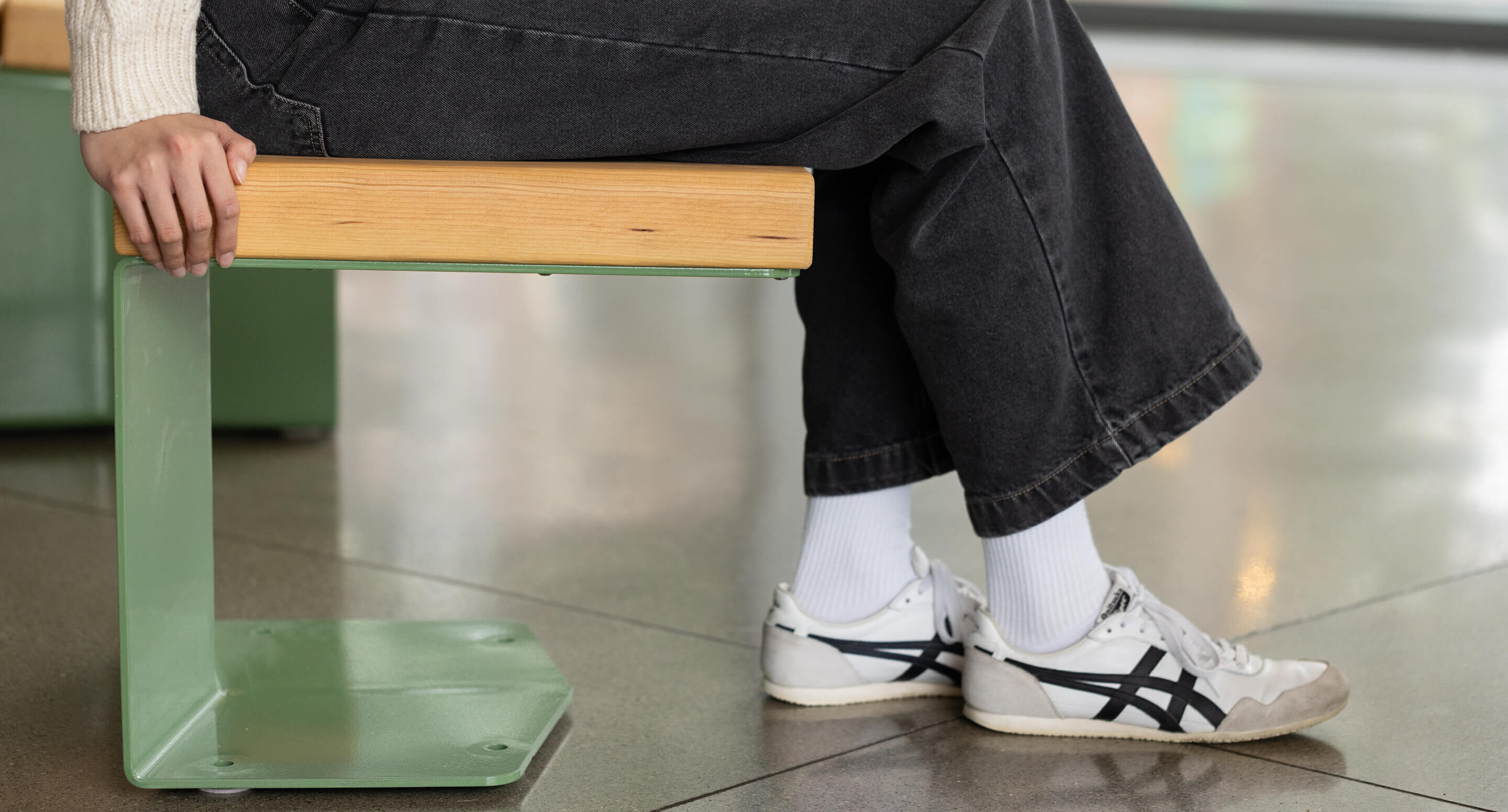 Personne assise sur un banc vert en métal et en bois, vêtue d'un pantalon noir, de chaussettes blanches et de baskets blanches à rayures noires.