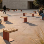 A modern outdoor plaza with spaced wooden benches; two people are seated and talking while others walk by in the background.