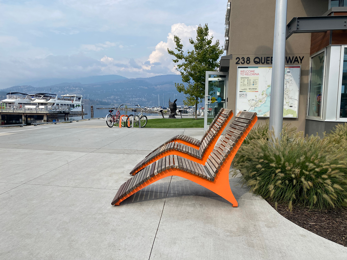 Two modern wooden lounge chairs with orange frames, designed for easy maintenance and care, are positioned on a sidewalk near a waterfront, with boats, bikes, and a map display in the background.