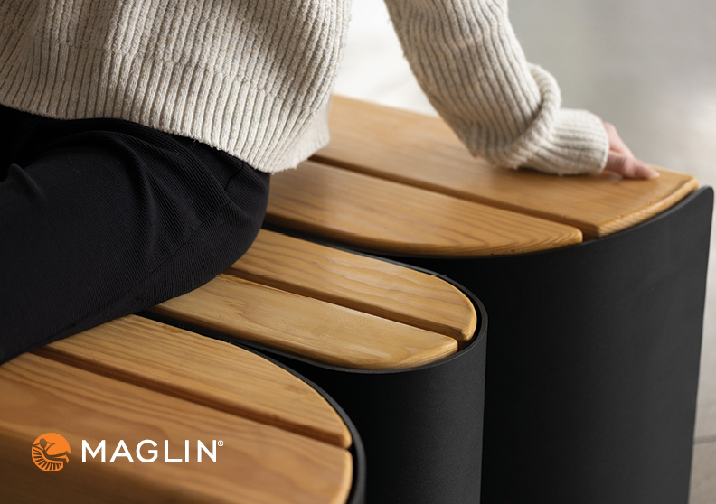 Person wearing a cream sweater and black pants sits on a modern wooden and metal bench. Maglin logo is visible in the lower left corner.