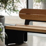 Banc en bois avec des supports noirs placé près de grandes fenêtres et d'une plante en pot, la lumière naturelle éclairant l'espace intérieur.