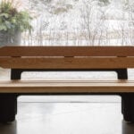 Un banc en bois avec un dossier et des pieds en métal noir est assis à l'intérieur près de grandes fenêtres en verre, avec une scène extérieure enneigée visible à l'arrière-plan.
