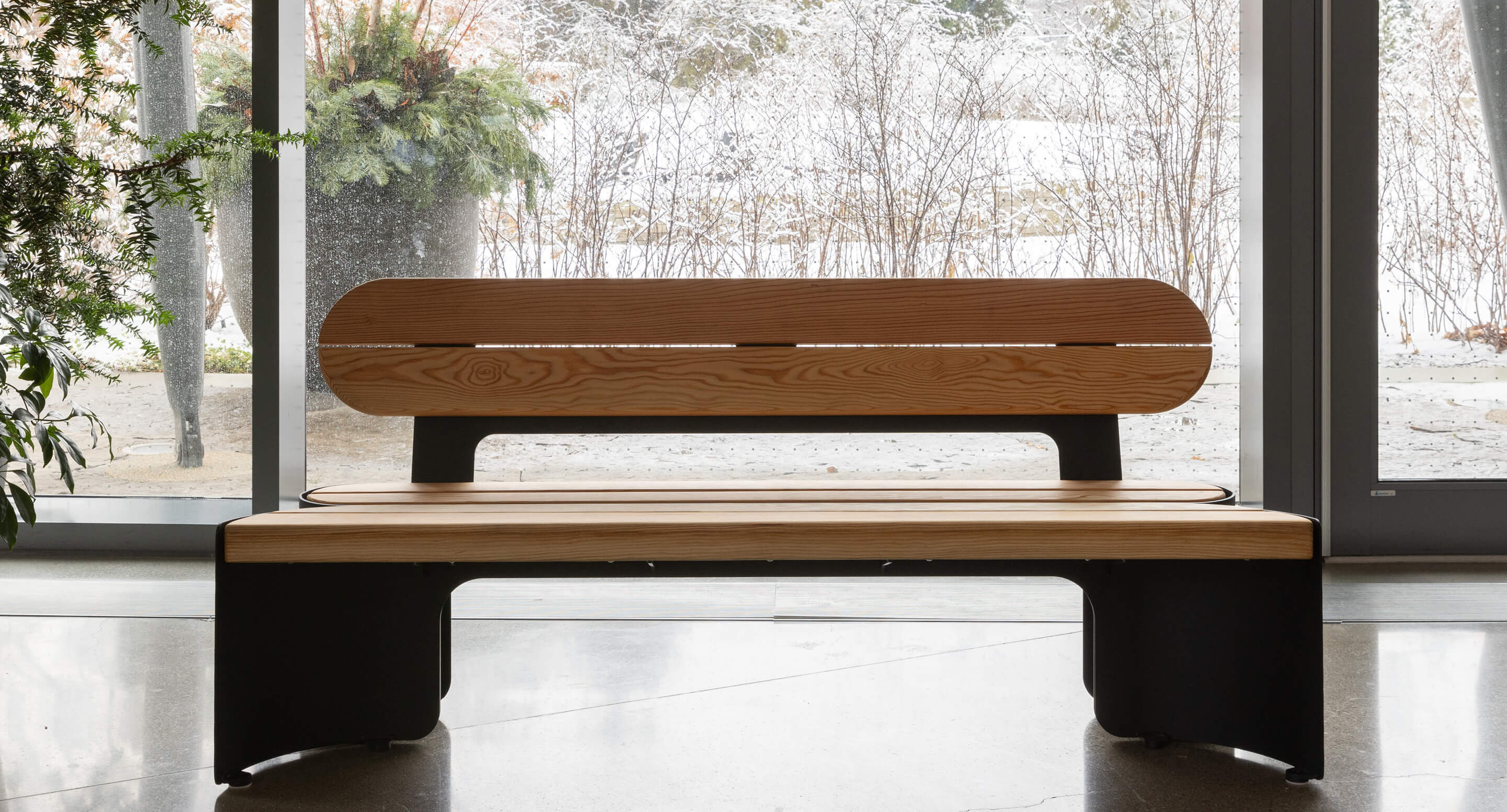 Un banc en bois avec un dossier et des pieds en métal noir est assis à l'intérieur près de grandes fenêtres en verre, avec une scène extérieure enneigée visible à l'arrière-plan.