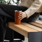 Personne assise sur un banc en bois, vêtue d'un pull crème et d'un jean noir, tenant un gobelet isotherme marron.